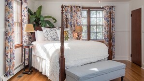 A four-poster bed with a white bedspread, two bedside lamps, a large potted plant, and a bench.