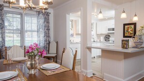 A dining area with a wooden table set for four, a vase with pink flowers, and a kitchen area with white cabinets and pendant lighting.