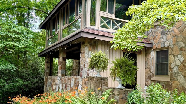 A stone house with a porch, surrounded by lush greenery and orange flowers.