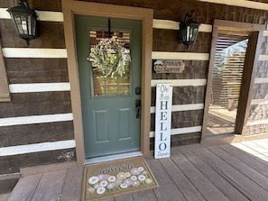 A wooden cabin entrance with a green door, a welcome mat, a 'Hello There' sign, and a 'Smokies Sunrise' sign.