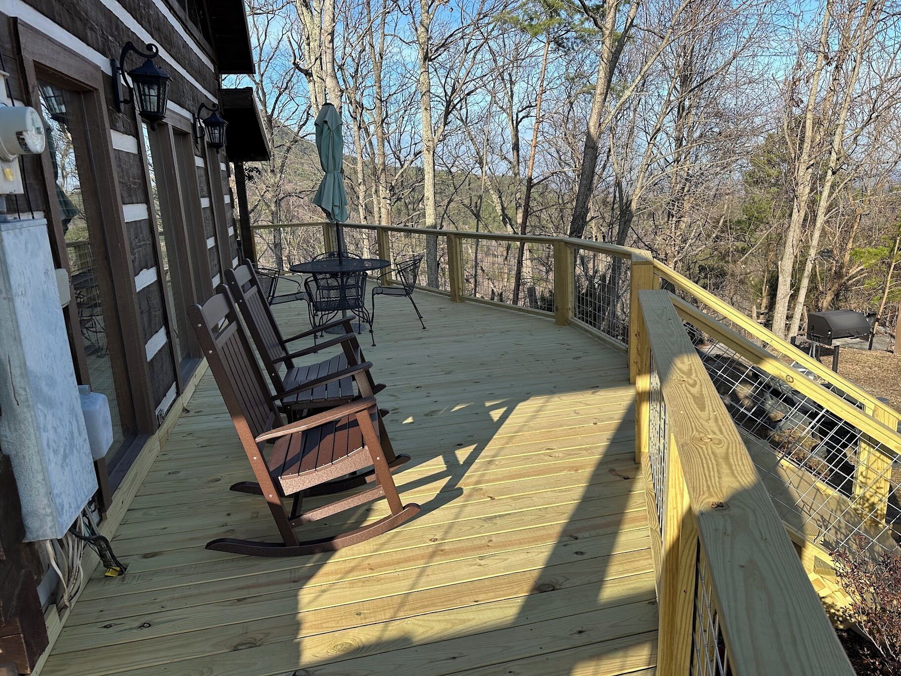 A wooden deck with a rocking chair and a table, surrounded by a wooden railing and trees.