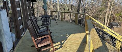 A wooden deck with a rocking chair and a table, surrounded by a wooden railing and trees.