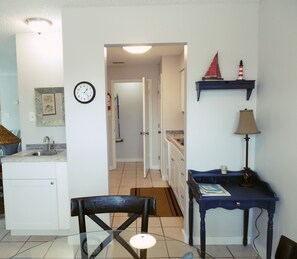 Een keuken met witte kasten, een spoelbak en een klok aan de muur. Er is een eettafel met stoelen en een blauwe tafellamp.