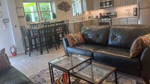 A modern kitchen with stainless steel appliances, a dining area with wooden chairs, and a living room with a leather couch and glass coffee table.