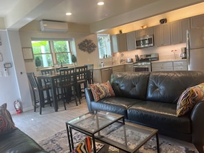 A modern kitchen with stainless steel appliances, a dining area with wooden chairs, and a living room with a leather couch and glass coffee table.
