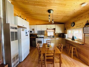 A wooden cabin interior with a kitchen, dining table, chairs, and a window with a view.