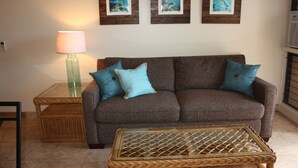Ein Wohnzimmer mit einem braunen Sofa, zwei türkisfarbenen Kissen, einem Korbtisch und einer Lampe auf einem Beistelltisch.