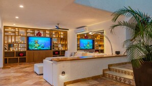 A modern living room with two large flat-screen TVs, a white sofa, a wooden entertainment unit, and a large potted plant. - Villa Luna Nueva - Concierge and Maid Service included (Cabo San Lucas)