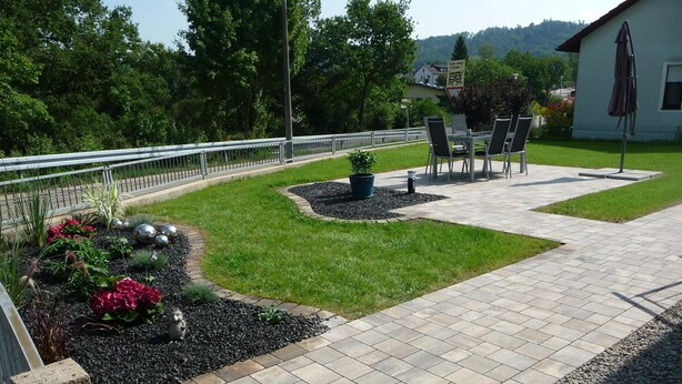 Un patio empedrado con una zona de estar, un pequeño jardín con flores y una planta en maceta, y un edificio al fondo.
