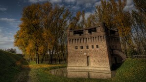 A historic stone tower with a fortified structure, surrounded by trees with autumn foliage, under a cloudy sky. - Holiday house by the river Po and surrounded by green in Emiliana (BONDENO)