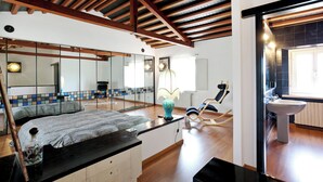 Un dormitorio con cama, mesita de noche, espejo, sillón mecedora y baño con lavamanos e inodoro.
