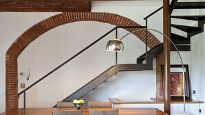 Un comedor con una mesa de madera, sillas y un cuenco de fruta. En el fondo, se ve una escalera con barandilla de metal y un arco de ladrillo.