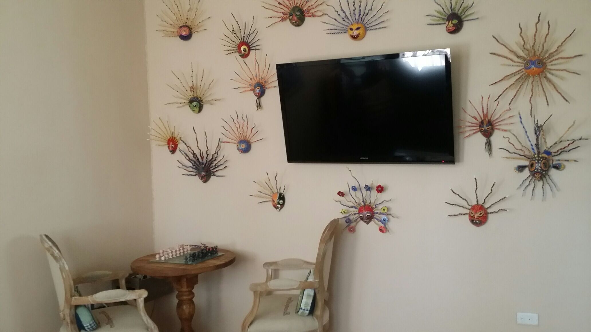 A room with a television mounted on the wall, surrounded by decorative wall art. There are two wooden chairs and a small round table with a chess set.