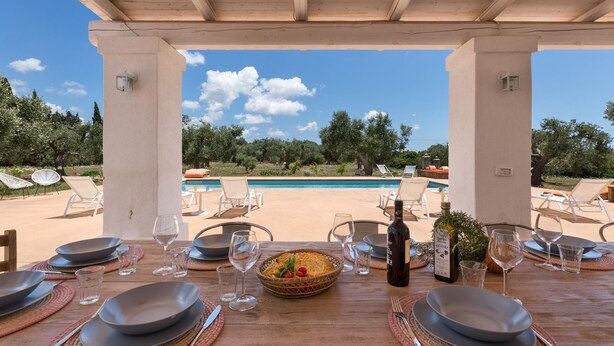Un área de comedor al aire libre con vista a la piscina, mesas dispuestas con platos, vasos y una botella de vino.