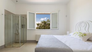 Un dormitorio con cama, ventana con vistas a árboles y zona de ducha con mampara de vidrio.