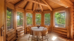 A wooden cabin with a round dining table and chairs, large windows, and a wooden ceiling.