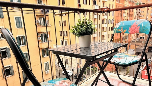 Een balkon met een tafel, twee stoelen, een bloempot en uitzicht op meerhoogbouw.