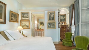 Una camera d'albergo con un letto grande, una poltrona verde, una cassettiera e quadri appesi alle pareti.