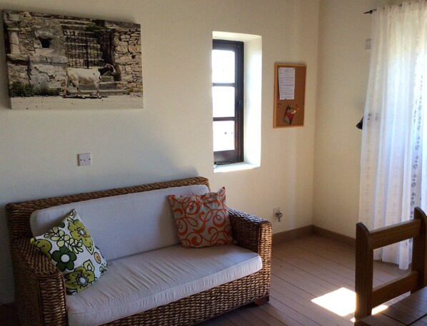 Una habitación con un sofá de mimbre, una ventana pequeña, un cuadro colgado en la pared y una silla de madera.