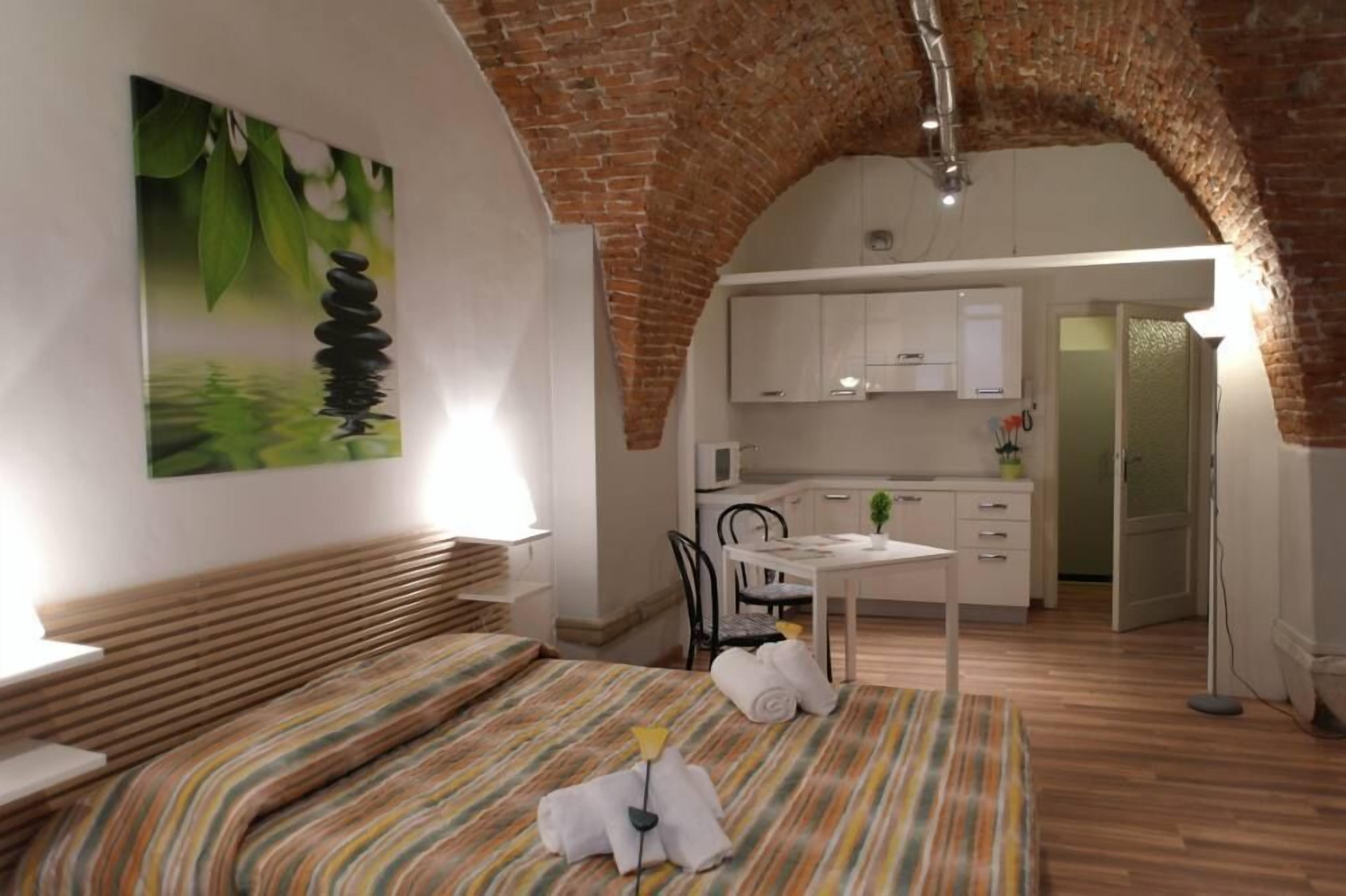 A bedroom with a bed, a painting, a dining area with a table and chairs, and a kitchen area with cabinets and a microwave.