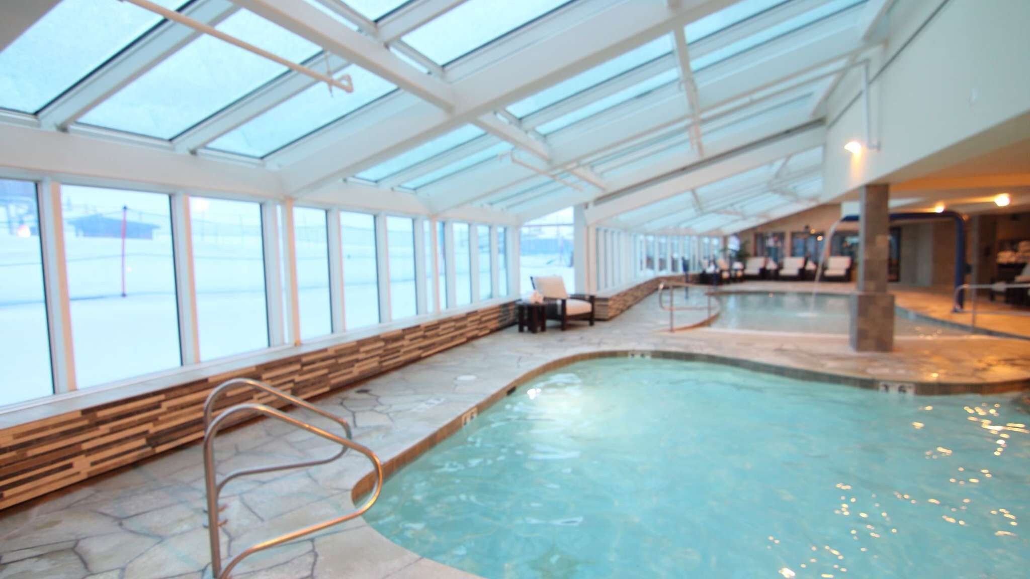 An indoor swimming pool with clear glass ceilings, stone flooring, and a wooden bench along the side.
