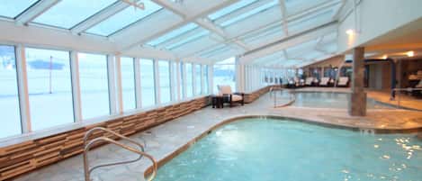An indoor swimming pool with clear glass ceilings, stone flooring, and a wooden bench along the side.