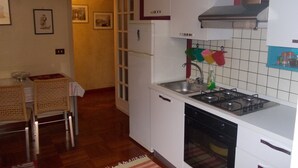 Kompakti keittiö, jossa on valkoiset kaapit, ruostumattomasta teräksestä valmistettu pesuallas, kaasuliesi ja astianpesukone. Keittiössä on puinen ruokapöytä tuoleineen, pieni matto ja ovi, joka johtaa toiseen huoneeseen.