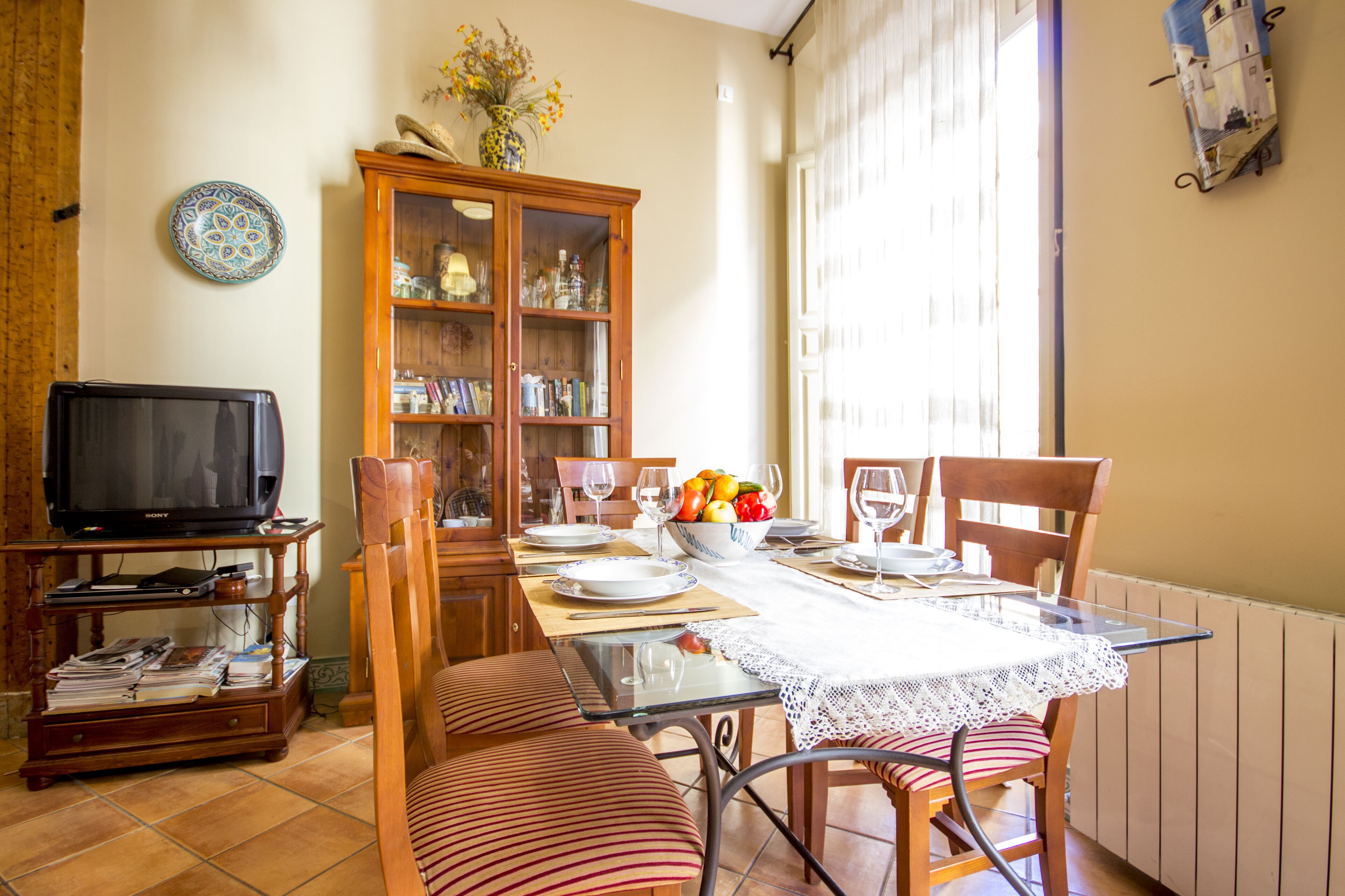 Un comedor con una mesa dispuesta para dos, un televisor y un aparador de madera con puertas de cristal.