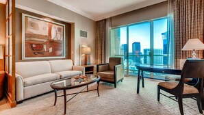 A modern hotel room with a large window offering a city view, a sofa, a coffee table, and a desk with a chair.