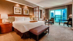 A hotel room with a large bed, a sitting area with a chair and table, and a view of the city through large windows.