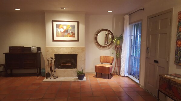 Una sala de estar con chimenea, un cuadro enmarcado, un espejo, un sillón de mimbre y una planta en maceta.