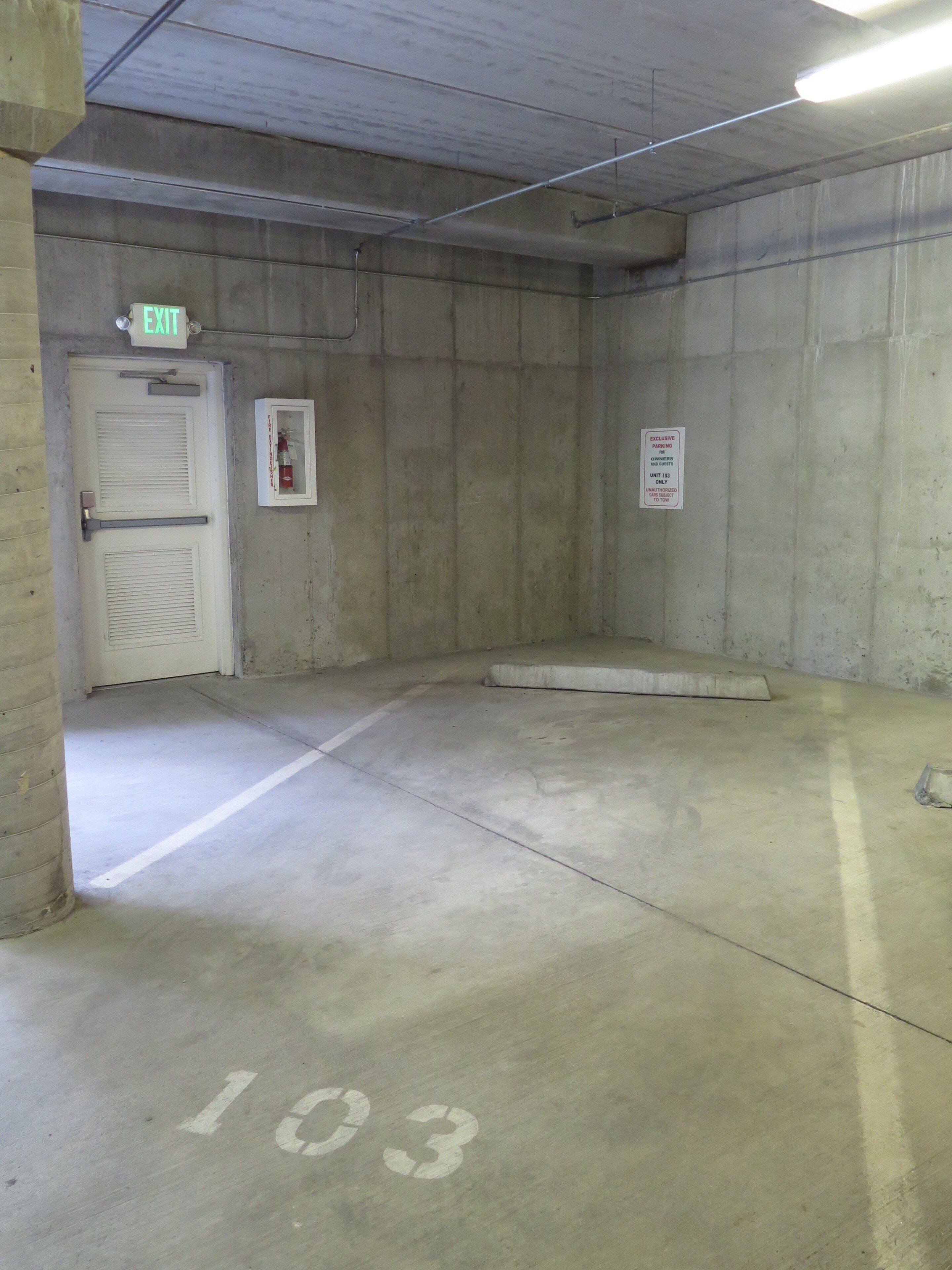 A parking garage with a concrete floor, marked parking space 103, an exit door, and a fire extinguisher.