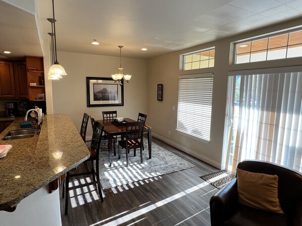 A modern kitchen with granite countertops, a dining area with a wooden table and chairs, and a large window with blinds.