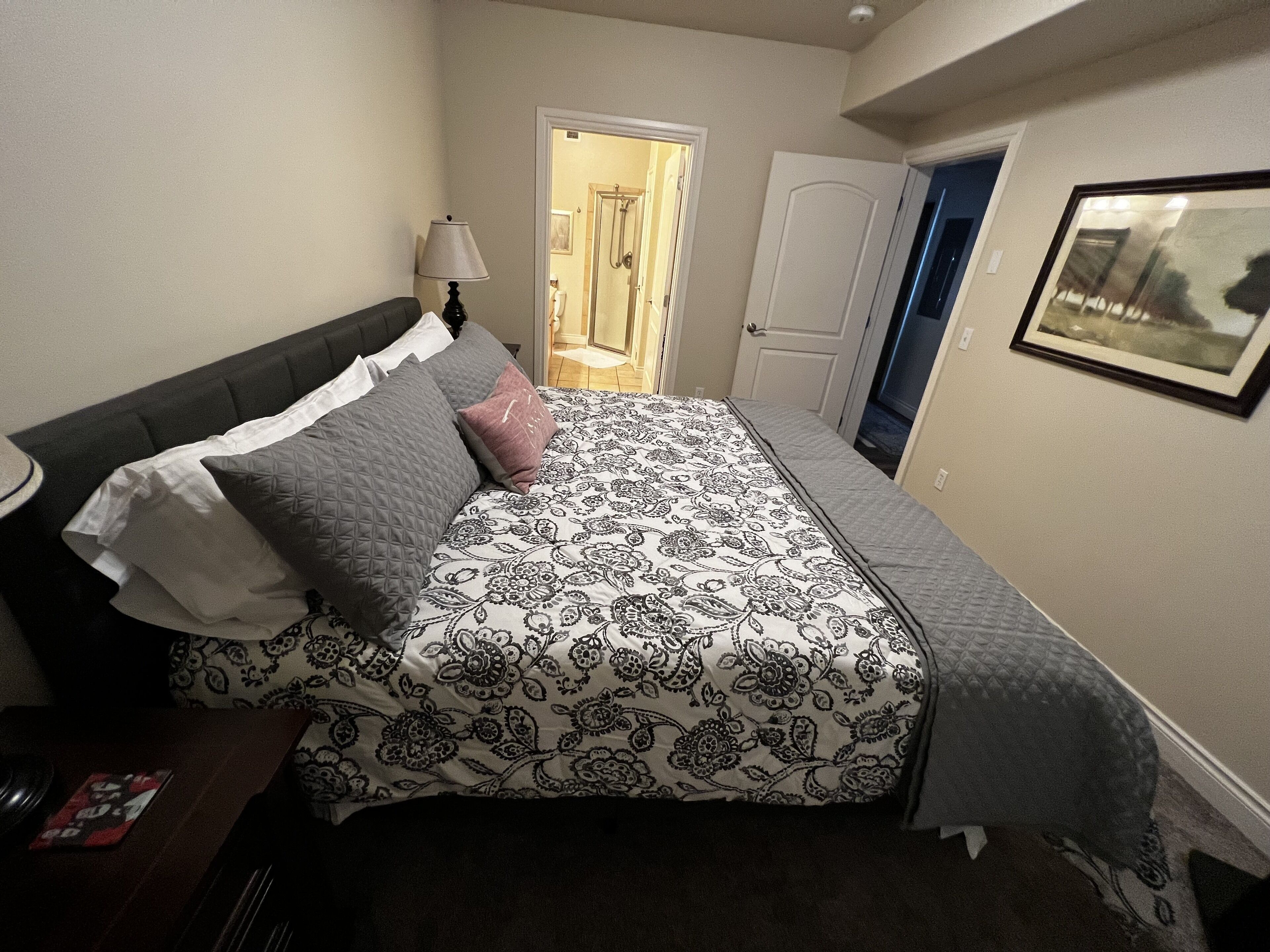 Un dormitorio con cama, mesita de noche, lámpara, puerta y un cuadro en la pared.