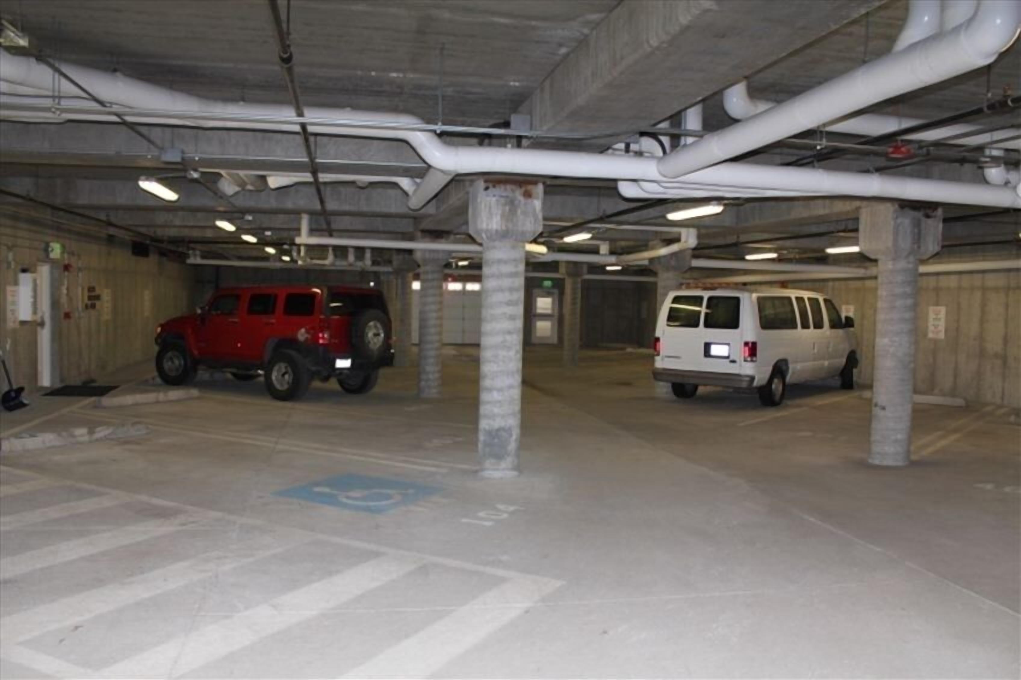 Un aparcamiento con dos vehículos estacionados, pilares de hormigón y tuberías aéreas.