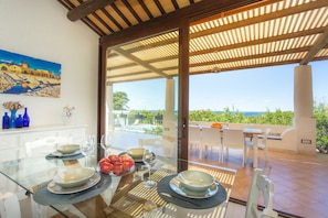 A dining area with a glass table, white chairs, and a view of the sea.