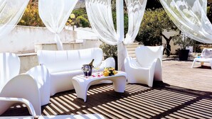 Un espace extérieur aménagé avec des meubles blancs, une table sur laquelle se trouvent une bouteille et des verres, et un décor de verdure en arrière-plan.