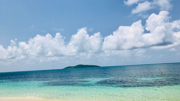 Ein Strand mit klarem türkisblauem Wasser, einem Sandufer und einer entfernten Insel.