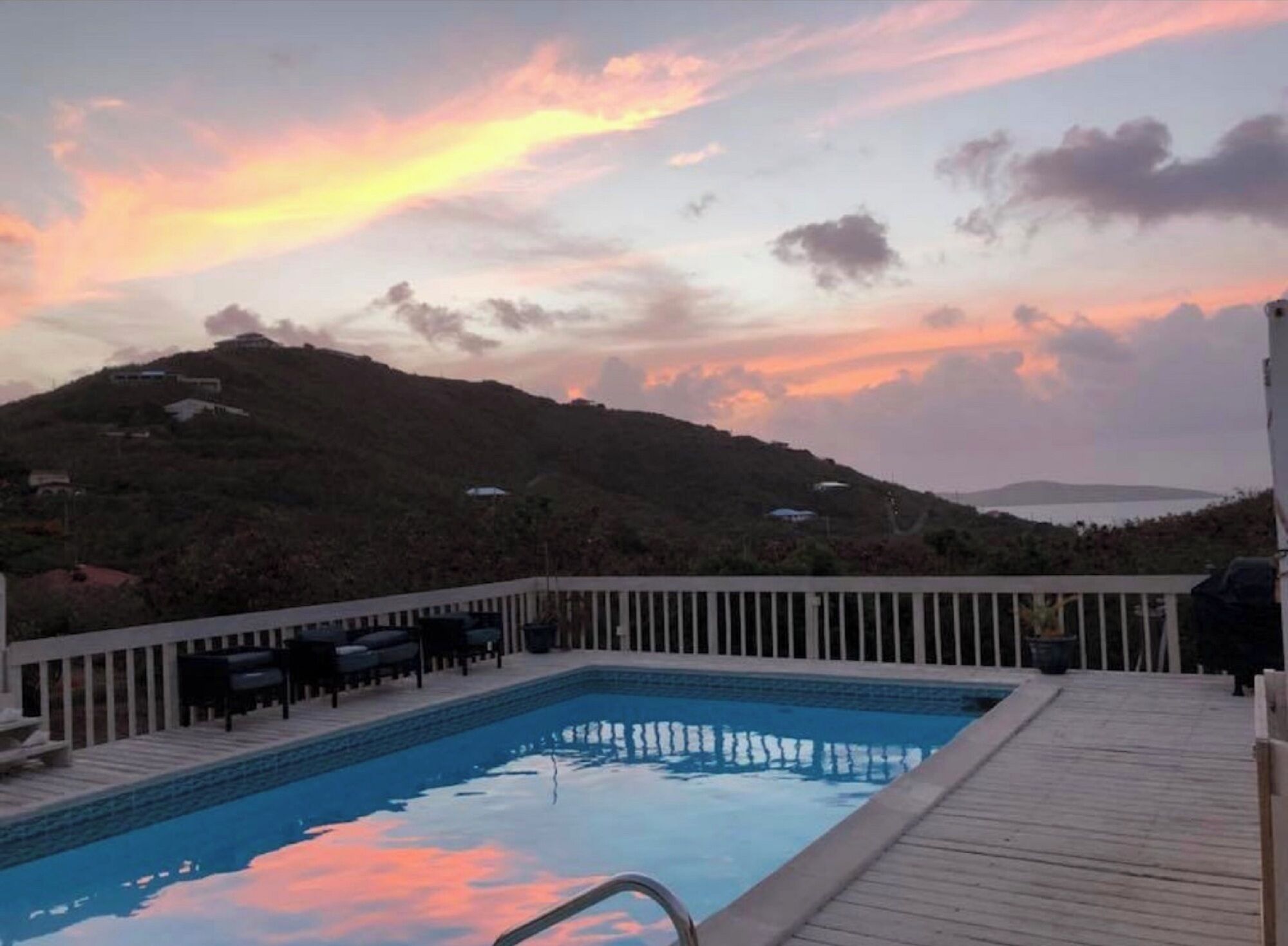 Piscina con muelle de madera y sillones de descanso, rodeada de colinas y un cielo al atardecer.