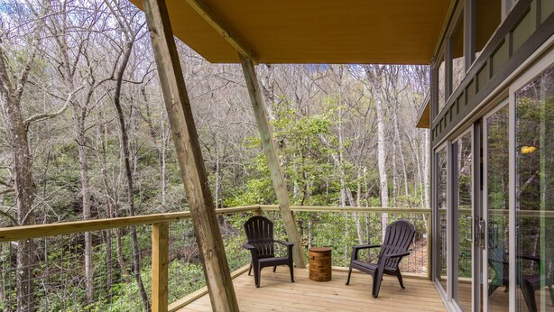 Eine Holzterrasse mit zwei Adirondack-Stühlen und einem kleinen runden Tisch mit Blick auf ein bewaldetes Gebiet.