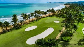 A golf course with sand bunkers and palm trees, overlooking a beach and ocean.