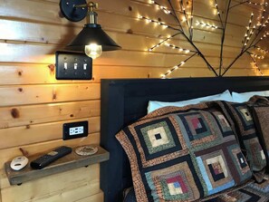 A cozy room with a wooden wall, a bed with a quilt, a lamp, and a wall-mounted power outlet.