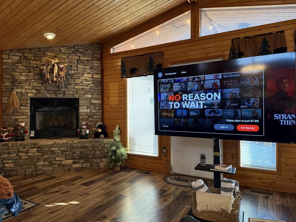 A cozy living room with a stone fireplace, a flat-screen TV displaying a streaming service, and a large window.
