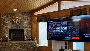 A cozy living room with a stone fireplace, a flat-screen TV displaying a streaming service, and a large window. - Honeymoon spot. Romantic. Great views. Private (Gatlinburg)