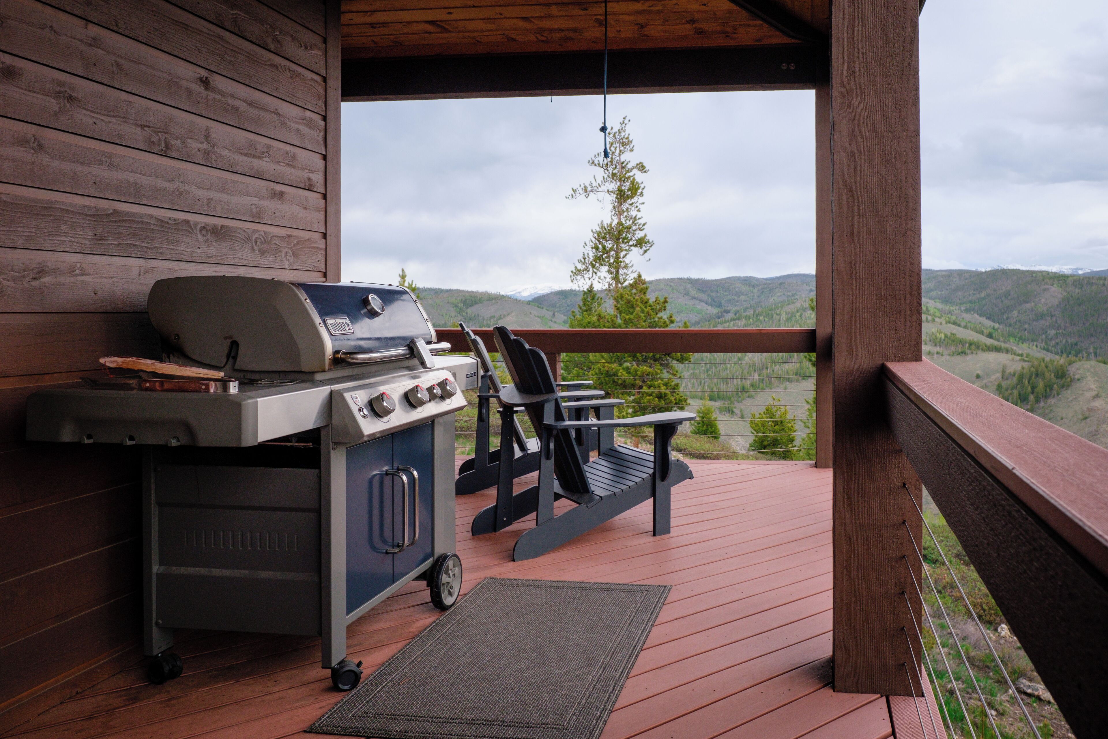 A covered outdoor area with a grill, a cart, and a chair, overlooking a mountainous landscape.