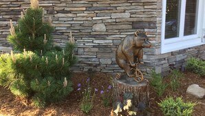 A stone wall with a bear sculpture on a tree stump, pine trees, and purple flowers.