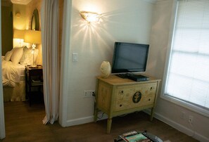 A bedroom with a bed, a bedside table with a lamp, a television on a wooden dresser, and a window with blinds.
