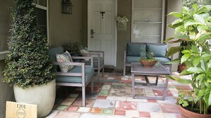 A cozy outdoor seating area with a wooden bench, a cushioned chair, and a table. There is a large potted plant on the left and another on the right. A door with a cross symbol is visible in the background. - Winter Park charm. Close to Park Ave (Winter Park)