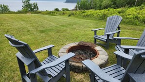 A fire pit with four Adirondack chairs in a grassy outdoor area with a view of a body of water and trees.