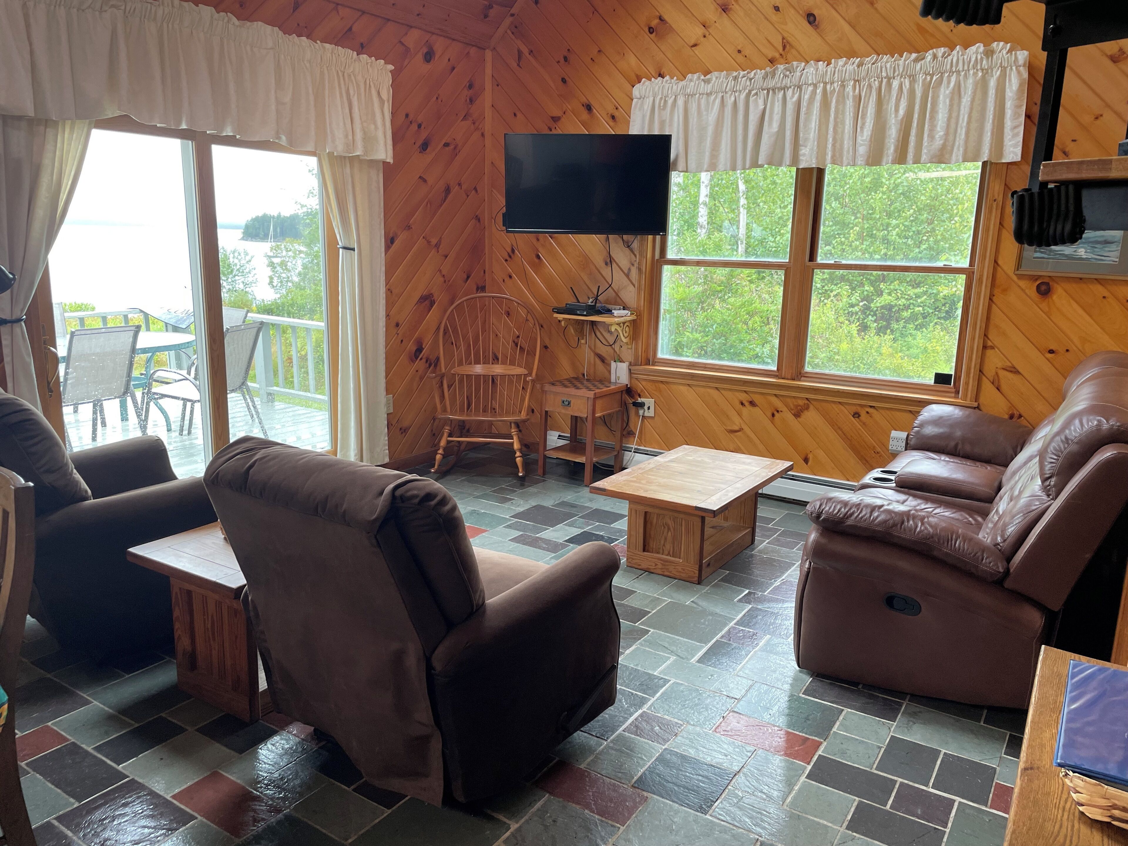 Interior acogedor de una cabaña con piso de madera, sofás de cuero marrón, una mesa de madera, un televisor montado en la pared y ventanas grandes con cortinas.